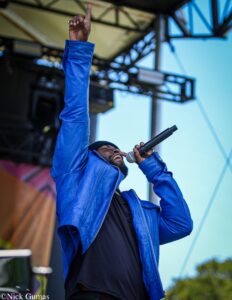A man in a blue jacket singing into a microphone on an outdoor stage, one hand raised with his index finger pointing upward.