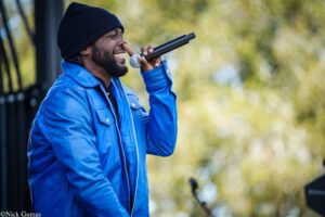 Man in a blue jacket and black beanie singing into a microphone outdoors, smiling with eyes closed.