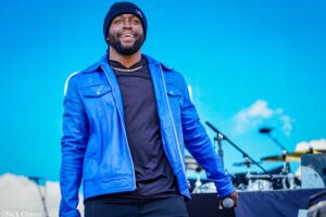 Male singer on a stage wearing a blue jacket and black beanie, holding a microphone and smiling with a bright blue background behind him.