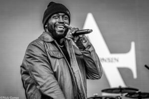 African American male singer wearing a beanie and leather jacket, smiling as he sings into a handheld microphone on stage.