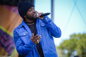 Man performing on stage, singing into a handheld microphone while wearing a blue jacket and dark beanie outside.