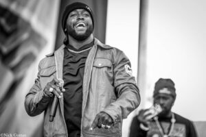 Black-and-white photo of a man wearing a beanie and jacket, singing into a microphone on stage, with another performer blurred in the background