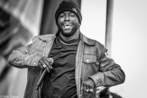 Male performer wearing a beanie and leather jacket, laughing while singing into a microphone on stage in black and white.