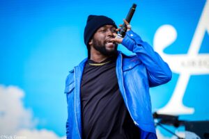 Male performer singing into a handheld microphone on stage, wearing a blue jacket and black beanie against a bright blue backdrop.
