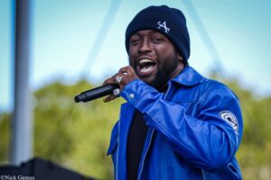 Man singing into a handheld microphone on an outdoor stage, wearing a blue jacket and dark beanie.
