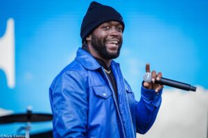 Male singer wearing a blue jacket and black beanie, smiling while holding a microphone on stage.