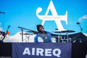 Male singer in a blue jacket and beanie performs on an outdoor stage with a large blue backdrop and white logo/text behind him.