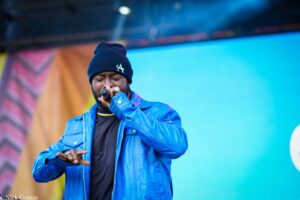 Male singer performing on stage, wearing a blue jacket and black beanie with an LA logo, holding a microphone close to his mouth.