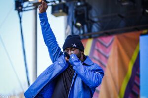 Singer on stage wearing a blue jacket and black beanie, raising a fist while singing into a microphone. (Informative, concise)