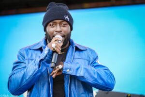 Male singer in a black beanie and blue jacket performing into a handheld microphone on a stage with a bright blue background.