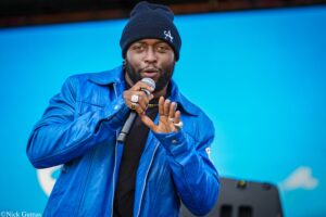 Male performer singing into a handheld microphone on stage, wearing a blue jacket and black beanie against a blue background.