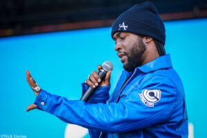 Man in a blue jacket and dark beanie with an LA logo sings into a handheld microphone on a stage, against a bright blue background.