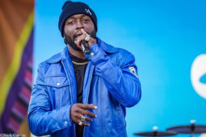 Male singer in a blue jacket and black beanie sings into a microphone on a bright blue stage backdrop.