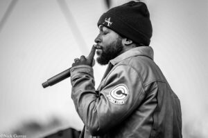 Black-and-white photo of a man wearing a beanie and jacket, holding a microphone near his mouth with a finger to his lips in a shushing gesture.