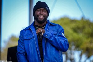 Smiling Black man in a blue jacket and black knit beanie, holding a microphone outdoors.