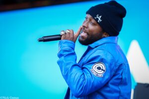 Male singer wearing a blue jacket and beanie, mic in hand, performing on stage against a bright blue background.