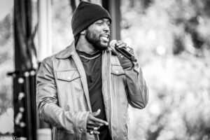 Black-and-white photo of a man singing into a handheld microphone on stage, wearing a beanie and jacket.