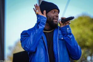 Male singer performing outdoors into a handheld microphone, wearing a blue jacket and black beanie.