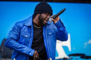 Man performing on stage, wearing a blue jacket and black beanie, singing into a handheld microphone with closed eyes.