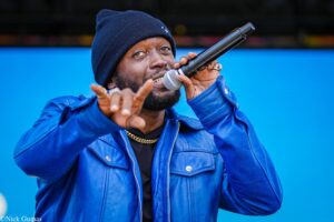 Male performer wearing a navy beanie and blue jacket singing into a handheld microphone, pointing toward the camera against a solid blue background.
