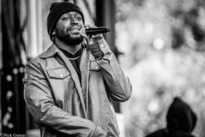 Man in a beanie and leather jacket sings into a handheld microphone, performing outdoors (black and white).