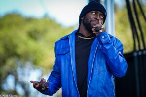Man in a bright blue jacket and black beanie gestures with his hands outdoors, wearing a gold chain and rings.