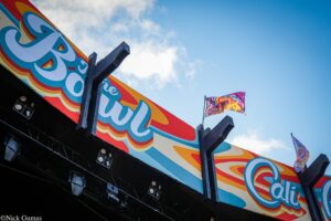 Colorful festival banner with swirling blue, orange, and red designs above a dark stage, against a blue sky with flags fluttering nearby.