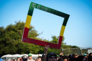 Colorful protest sign reading 'CONNECTING' held above a crowd at an outdoor rally, with green/yellow/red vertical supports against a blue sky.