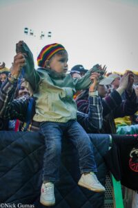 Child sits on an adult's shoulders at a crowded outdoor event, wearing a rainbow striped hat and green jacket.