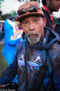 Close-up of a man wearing a patterned cap and galaxy-print hoodie, holding a Nikon camera at an outdoor event.