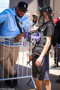 Two men at a street event by a metal barrier; one in a blue striped shirt and LA cap leans over, the other with a long gray beard and a black outfit standing beside a purple device.