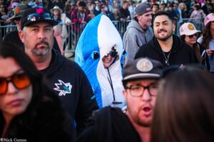 Crowd at an outdoor event; a person wearing a blue shark hoodie smiles among attendees in casual clothing.