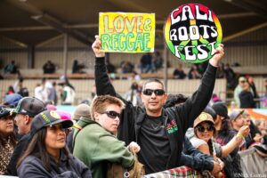 Crowd at a festival holding colorful signs: a yellow sign reading 'LOVE & REGGAE' and a round rainbow sign saying 'CAL ROOTS FEST' with people cheering in the background.