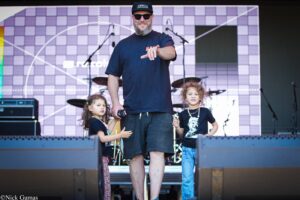 Stage scene: a man in a dark shirt and cap points at the camera, with two children beside him, drums and equipment in the background behind a grid backdrop.