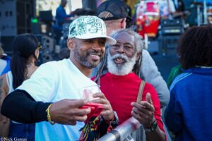 Two men at an outdoor event posing for a photo, one in a white cap with a floral design smiling at the camera, holding a red device and a cup.