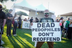 Person wearing a clown mask and cap holds a large protest sign outdoors, with a crowd and tents in the background.