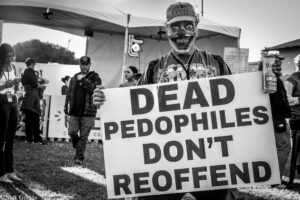 Protester wearing a clown mask and cap holds a large banner with a bold anti-pedophile message at an outdoor event.