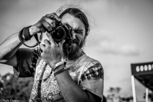 Black-and-white photo of a bearded man aiming a Nikon camera toward the viewer, capturing a shot outdoors.