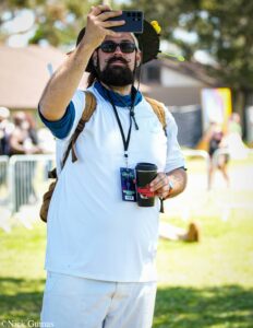 Man in a white outfit and hat takes a selfie with a smartphone outdoors, wearing sunglasses and a decorative mustache, holding a coffee cup.
