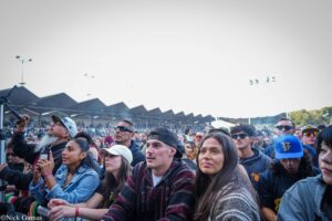 Crowd of diverse people at a daytime outdoor concert, close to the stage with barriers in the foreground.