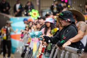 Person in a straw hat and black hoodie aiming a bubble gun at a cheering crowd behind a barrier, bubbles filling the air.