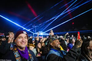 Close-up of a smiling man in a maroon beanie at a crowded night concert with blue laser lights in the background.
