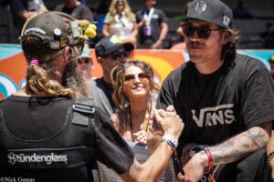 Two men shake hands at an outdoor event; foreground man wears a black Vans shirt and tattooed arms, onlookers behind him.