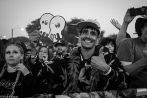 Smiling man in a cap holds a paper ghost cutout at a crowded outdoor parade while others cheer in the background.