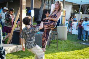 A man in camouflage shirt lifts a woman in a tiger-print dress during an outdoor festival.