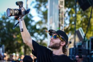 Male photographer wearing a cap and sunglasses holds a professional video camera on a monopod at an outdoor event with a blurred crowd in the background
