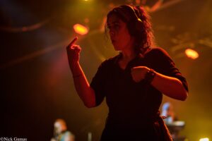 Female dancer on stage wearing headphones, illuminated by warm orange stage lights in a dark venue.