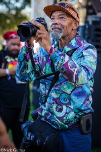 Photographer in a vibrant patterned jacket aims his Nikon camera, outdoors, smiling under a brimmed hat with sunglasses tucked in.