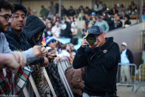 Photographer wearing a Kona cap and black jacket takes a photo of fans behind a barrier at an outdoor event.
