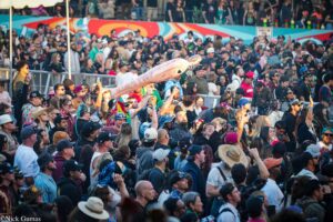 Dense crowd at an outdoor festival, with banners and a long pale inflatable being waved above heads against a colorful mural backdrop.